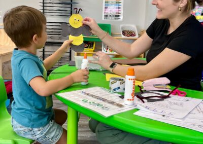 A photo of one of our occupational therapists, Miss Danielle, sitting at a table with a child working on a craft. Danielle and the student both hold up their finished product which is a bumble bee.