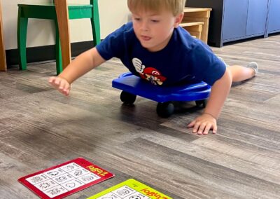 A child with blond hair lays on his stomach on a scooter and crawls across the floor to reach the game he is playing.