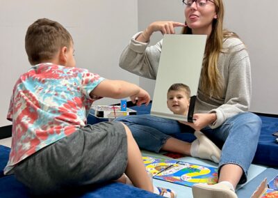 A photo of one of our speech therapists, Micc Rachel, and a child she is working with in speech therapy. Rachel is holding up a mirror and pointing to her tongue as the child sees himself in the mirror to practice saying the same sound.