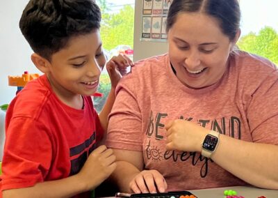 A photo of one of our occupational therapists sitting at a table with a child. The two of them are next to each other as they play a strategy game. Both of them have big smiles on their face.