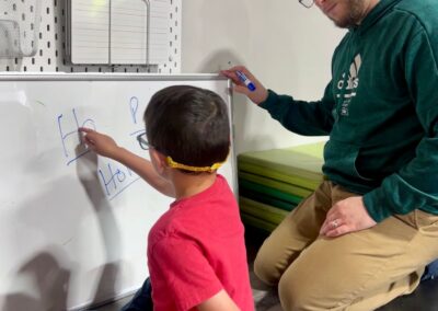 A photo of one of our lead therapists, Mr Mike, working with a child to write words on a whiteboard and work on pronunciation. The child is in a red shirt and sitting on the ground pointing at the whiteboard. Mr Mike is watching the child attentively.