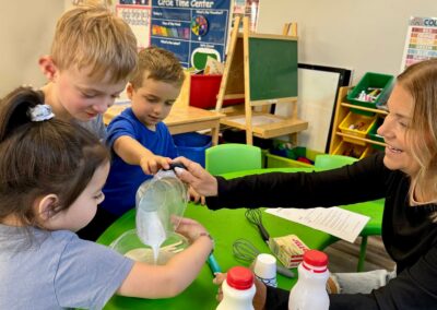 This photo shows Miss Stacey, our preschool teacher, making popsicles with her preschool students. The kids are helping pour a measuring cup of milk into a bowl. Everyone is smiling and watching attentively.